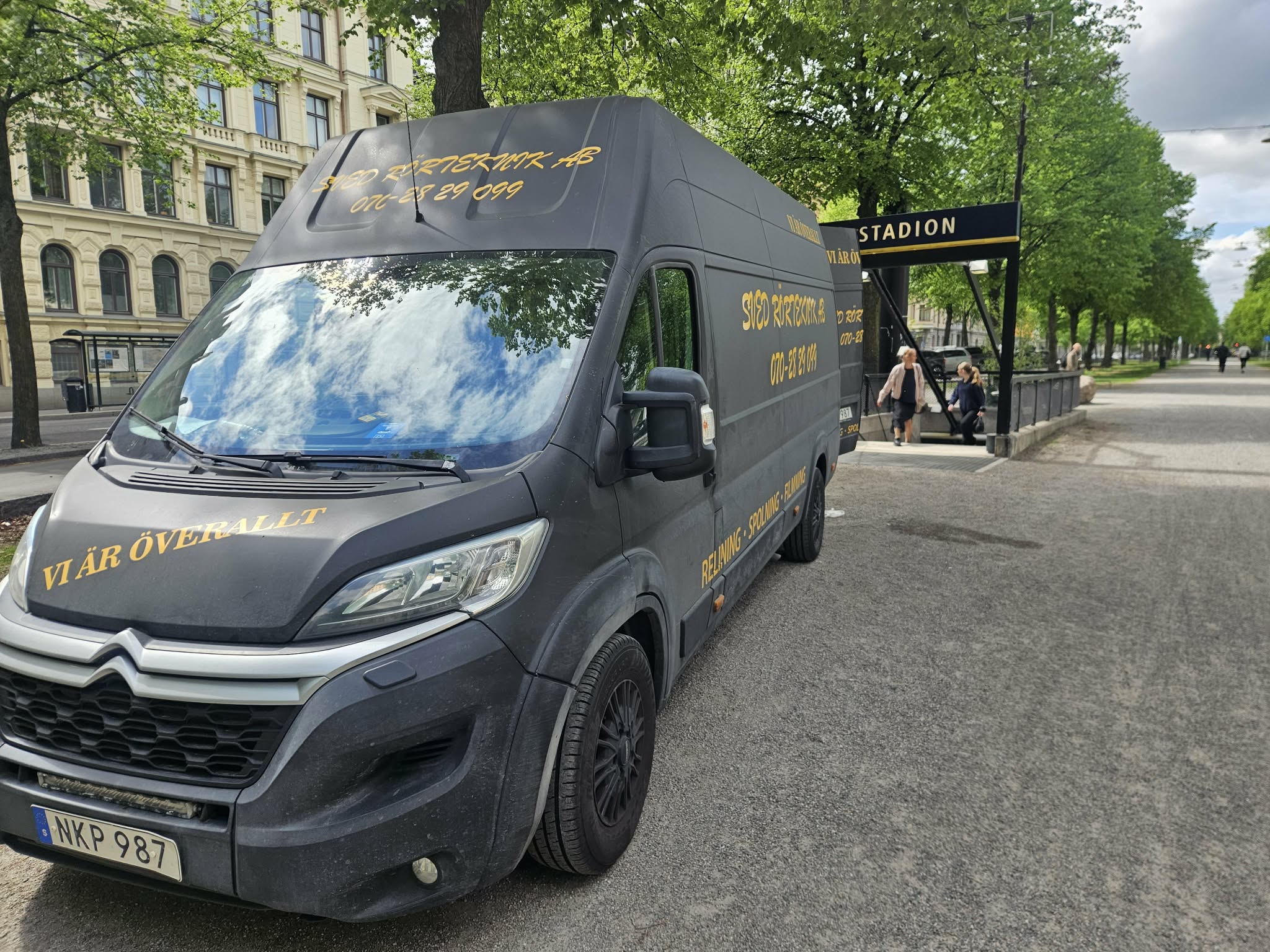 Servicebilen sedd från sidan på en gata med allé och kringliggande bebyggelse.
